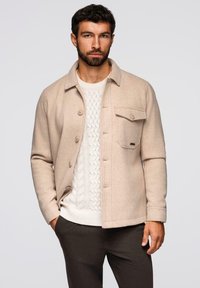 Giacca in misto lana beige con chiusura a bottoni, taschino singolo sul petto e una texture sottile, indossata sopra un maglione bianco a trecce.