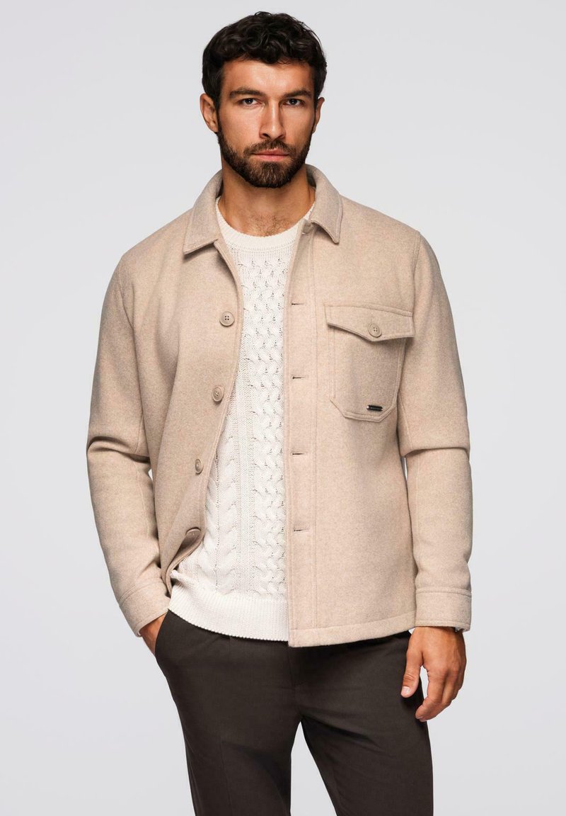 Giacca in misto lana beige con chiusura a bottoni, taschino singolo sul petto e una texture sottile, indossata sopra un maglione bianco a trecce.