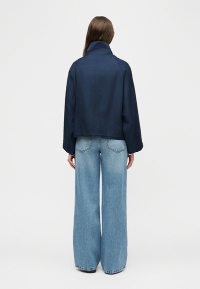 Γυναίκα που φοράει μπλε σκούρο σακάκι και ανοιχτό μπλε παντελόνι με φαρδιές παντελόνες, στέκεται με την πλάτη στην κάμερα σε ένα απλό φόντο.