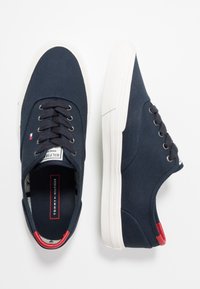 Marineblaue Canvas-Sneaker mit weißen Gummisohlen, runden Zehen, sechs Ösen und roten Akzenten an Ferse und Zunge. Leicht strukturierte Oberfläche.