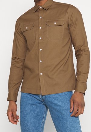 Mand iført en brun skjorte med knapper og to forlommer, der står mod en ensfarvet baggrund med blå jeans.