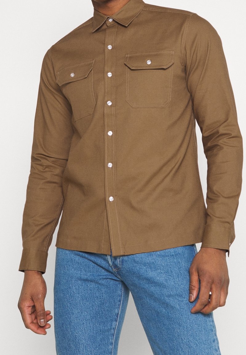 Homme portant une chemise marron à boutons avec deux poches à rabat sur le devant, associée à un jean bleu, debout devant un fond uni.