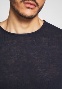 Detailní záběr mužské dolní části obličeje a krku, který má na sobě tmavě modrý volně pletený svetr s kulatým výstřihem.