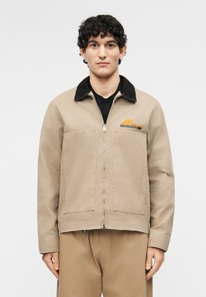 Νεαρός άνδρας με κοντά μπούκλια, φοράει μπεζ μπουφάν με φερμουάρ και μαύρο γιακά, συνδυασμένο με μπεζ παντελόνι, στο φόντο λευκού χρώματος.