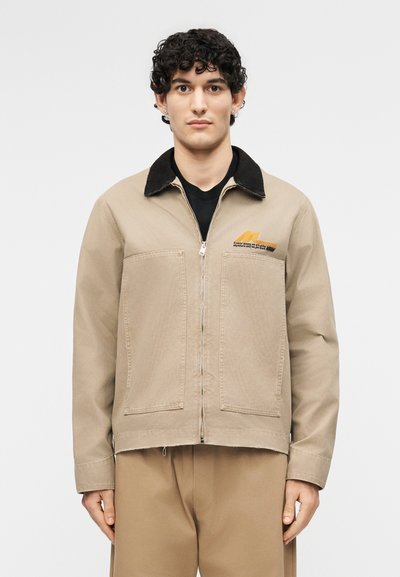 Jeune homme aux cheveux courts et bouclés portant une veste beige à fermeture éclair avec col noir et un pantalon beige assorti sur fond blanc.