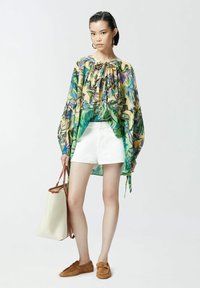 Lagani, bluzasti kroj s cvjetnim uzorkom i mašnom sprijeda, uparen s bijelim kratkim hlačama i smešnim mokasinkama. Drži kremastu torbu s smešnim akcentima.