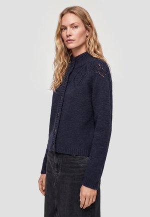 Blonde vrouw draagt een donkerblauwe gebreide cardigan met knopen, een textuurbreiwerk en een hoge hals, gecombineerd met een zwarte rok tegen een effen achtergrond.
