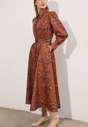 Kobieta w rdzawo-pomarańczowej sukience midi z ciemnym kwiatowym wzorem, z długimi rękawami i dopasowaną talią, stojąca na tle białej ściany.