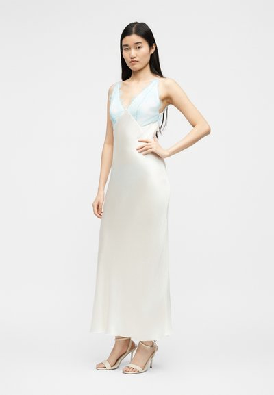 Νεαρή γυναίκα στέκεται με το χέρι στη μέση, φορώντας ένα αμάνικο, λευκό σατέν φόρεμα με V λαιμόκοψη, λεπτομέρειες από ανοιχτό μπλε δαντέλα και μπεζ ψηλοτάκουνα σανδάλια.