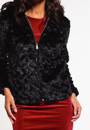 Femme portant une veste zippée noire texturée sur une robe en velours rouge, les mains reposant le long du corps.