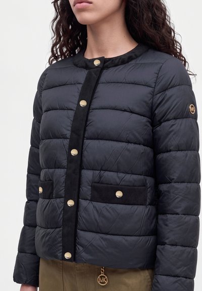 Modèle portant une veste matelassée foncée avec des boutons dorés, des bordures noires, deux poches et un pendentif orné du logo.