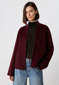 Giacca borgogna con zip frontale e vestibilità rilassata. Presenta due tasche frontali e colletto a costine. Abbinata a jeans a gamba larga blu chiaro.