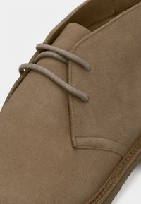 Κοντινή λήψη καφέ σουέτ μποτάκι ερήμου με μπεζ κορδόνια και εμφανή ραφή κατά μήκος της σόλας και του άνω μέρους.