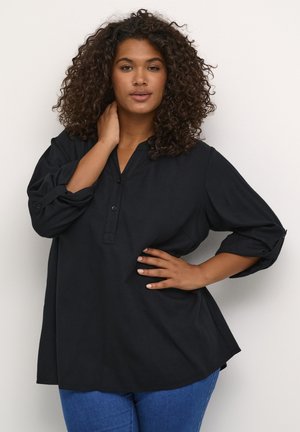 Chemise noire à boutons, avec col, manches trois-quarts et coupe décontractée. Le tissu a une texture douce. Portée avec un jean bleu.