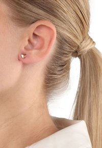 Närbild av en blond kvinna som bär en liten, glänsande, roseguld hjärtformad studörhänge, med håret uppsatt i en hästsvans.