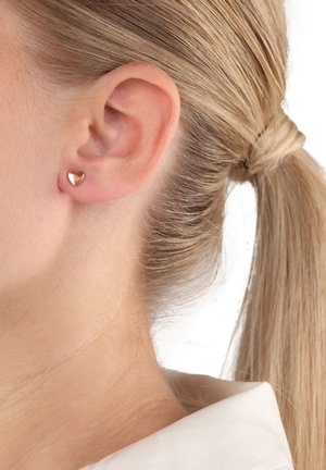 Primo piano di un orecchio di una donna bionda che indossa un piccolo orecchino a forma di cuore in oro rosa lucido, con i capelli raccolti in una coda di cavallo.