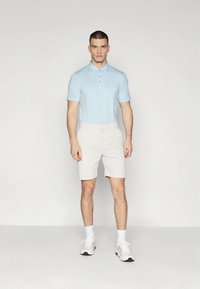 Világoskék galléros póló krémszínű rövidnadrággal párosítva. Az egyén fehér tornacipőt visel és rövid hajat, egyenes testtartással áll egyszerű háttér előtt.