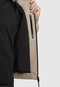 Giacca zip-up con esterno beige, interno nero e tasca con zip visibile. Texture morbida e vita elastica nella parte inferiore.