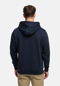 Laivastonsininen huppari, jossa on kiristettävä huppu ja resorit hihansuissa, valmistettu pehmeästä ja teksturoidusta kankaasta. Edessä on kengurutasku.