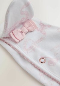 Bílá látka s jemnými růžovými vzory má růžovou mašli, dekorativní prošívání a akcent z růžového zlata ve tvaru kroužku. Měkká textura a zaoblený tvar.