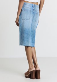 Femme portant une jupe en denim bleu clair arrivant aux genoux avec un ourlet effiloché et des sandales à plateforme à talons hauts marron, se tenant sur un fond uni.