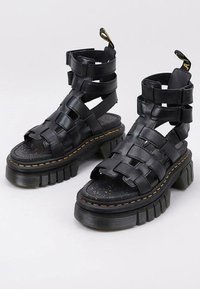 Sandalias de plataforma negras con tiras, suela gruesa, diseño de cuero trenzado y plantilla moteada. Presenta un detalle de costura amarilla distintiva.