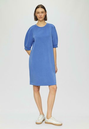 Blaues, knielanges Kleid mit kurzen Puffärmeln, aus glattem Stoff. Kombiniert mit weißen Sneakern mit beigem Sohlen.