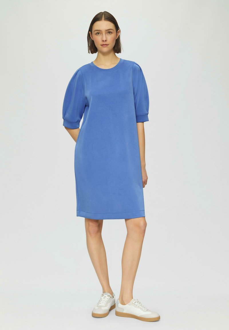 Blaues, knielanges Kleid mit kurzen Puffärmeln, aus glattem Stoff. Kombiniert mit weißen Sneakern mit beigem Sohlen.