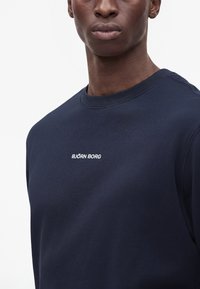 Homme portant un sweat-shirt bleu marine à col rond avec un petit texte blanc "BJÖRN BORG" sur la poitrine, sur un fond blanc uni.