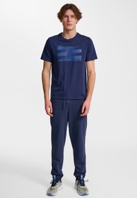 Marinblå t-shirt med kort ärm med blå text och logotyp "PERFORMANCE", matchad med marinblå joggerbyxor och grå sneakers med blå detaljer.