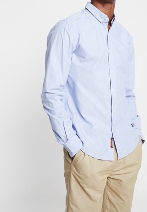 Mand iført en lyseblå skjorte med knapper og beige bukser, hænderne i lommerne, stående mod en ensfarvet hvid baggrund.