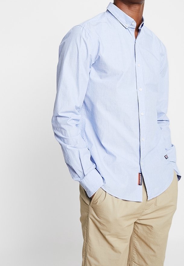 Homme portant une chemise bleu clair boutonnée avec un pantalon beige, mains dans les poches, debout devant un fond blanc uni.