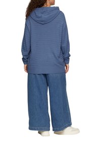 Blauwe hoodie met structuurstreepdesign, gecombineerd met wijde denim broek. Model staat met de rug naar de camera gekeerd.