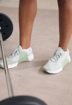 Personne portant des baskets Nike gris clair avec lacets debout près d'une barre avec des poids sur le sol d'une salle de sport.