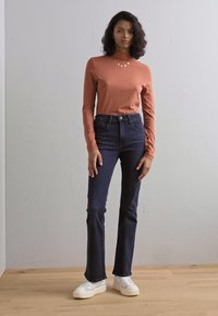 Langemouwige rustkleurige T-shirt met een hoge hals, gecombineerd met donkerblauwe flared jeans en witte sneakers op een lichte houten vloer.