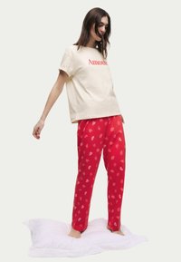 Katoenen T-shirt in crème met rode tekst "Amour", gecombineerd met rode pyamabroek met een lippenafdruk in lichtere tinten, staande op een wit kussen.