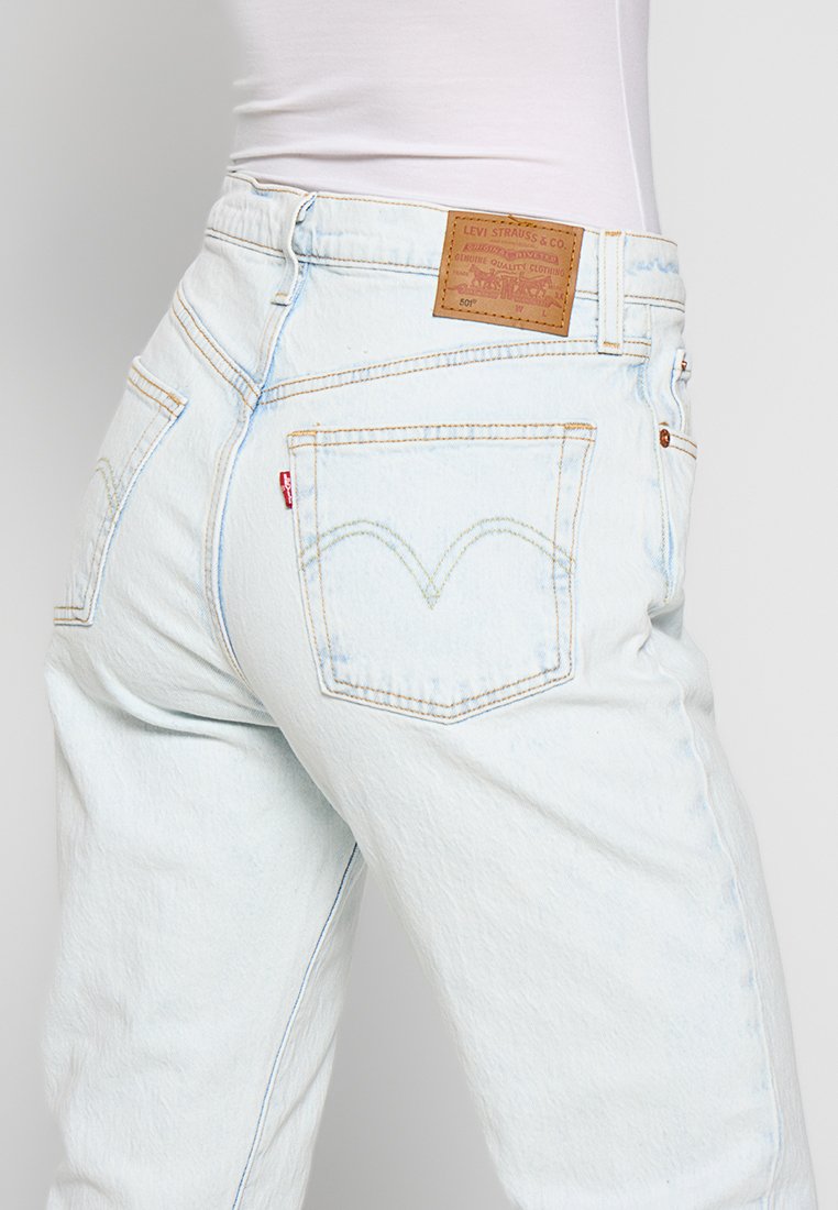 Jeans de mezclilla en un azul claro con cintura alta, que presentan un parche de cuero marrón y un detalle de costura icónica en el bolsillo trasero.