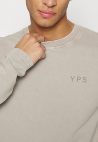Camiseta de manga larga beige con cuello redondo, puños acanalados y pequeño logotipo "YPS" en un color a juego en el pecho. Textura de tela suave.