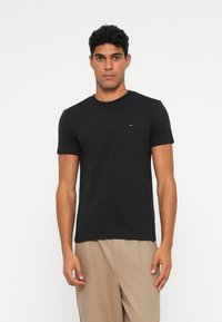 Tricou negru din bumbac cu guler rotund, mâneci scurte și un mic accent cu logo pe piept, purtat cu pantaloni bej deschis.