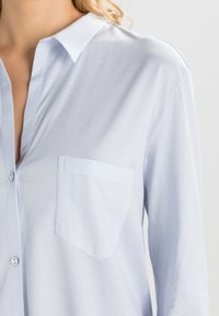 Jasnoniebieska koszula z guzikiem, wykonana z miękkiego materiału, z klasycznym kołnierzykiem, kieszenią na piersi oraz długimi rękawami o luźnym kroju.