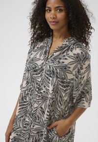 Abito camicia stampato in beige chiaro con motivo a foglie nere, chiusura frontale con bottoni, maniche corte e tasche laterali per una funzionalità aggiuntiva.