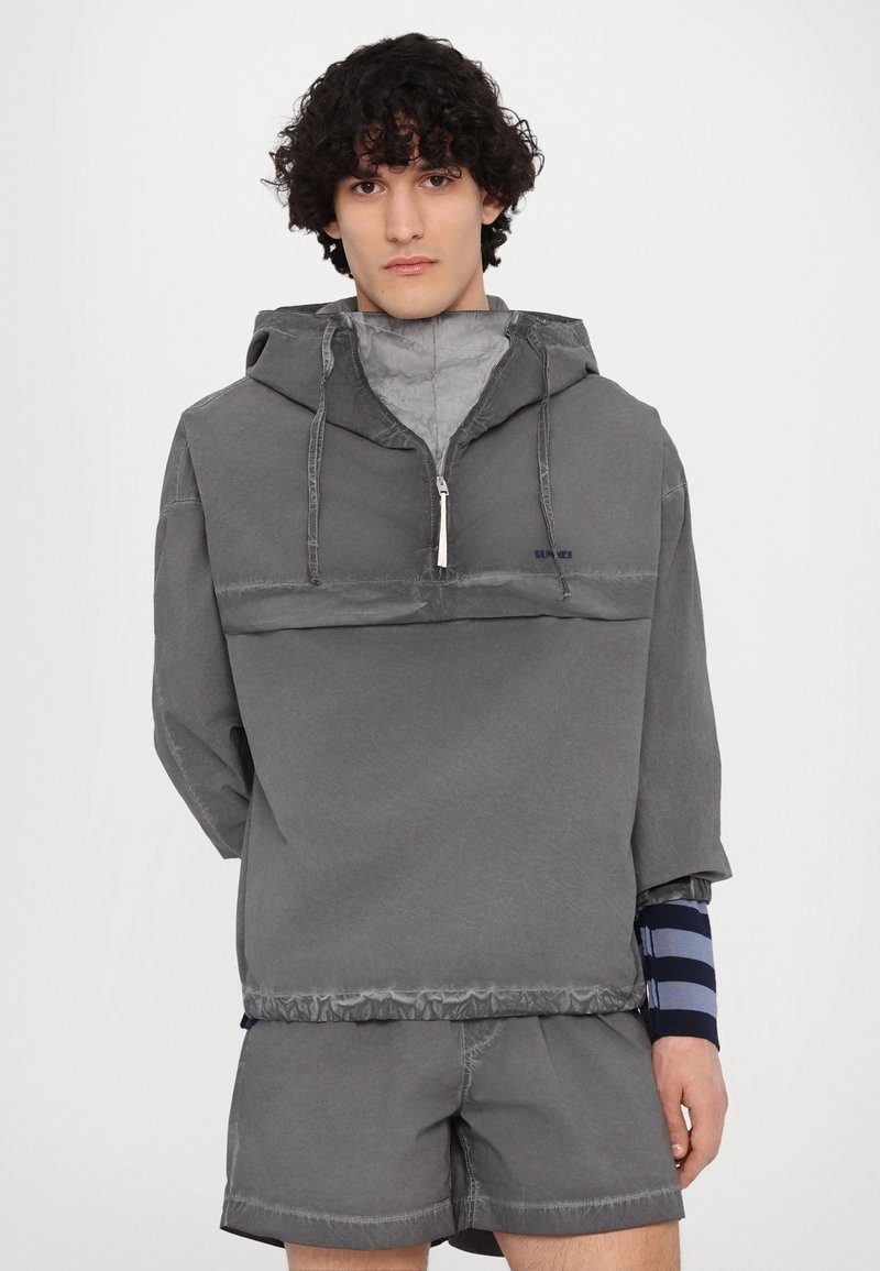 Pullover grigio con zip a metà davanti e cappuccio, realizzato in un tessuto testurizzato. Include anche pantaloni corti grigi abbinati con dettagli di cucitura visibili.