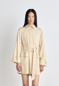 Béžové šaty s opaskom, golierom, dlhými rukávmi a prednými vreckami. Tkanina pôsobí hladko s miernym leskom. Jednoduchý dizajn a prispôsobený strih.