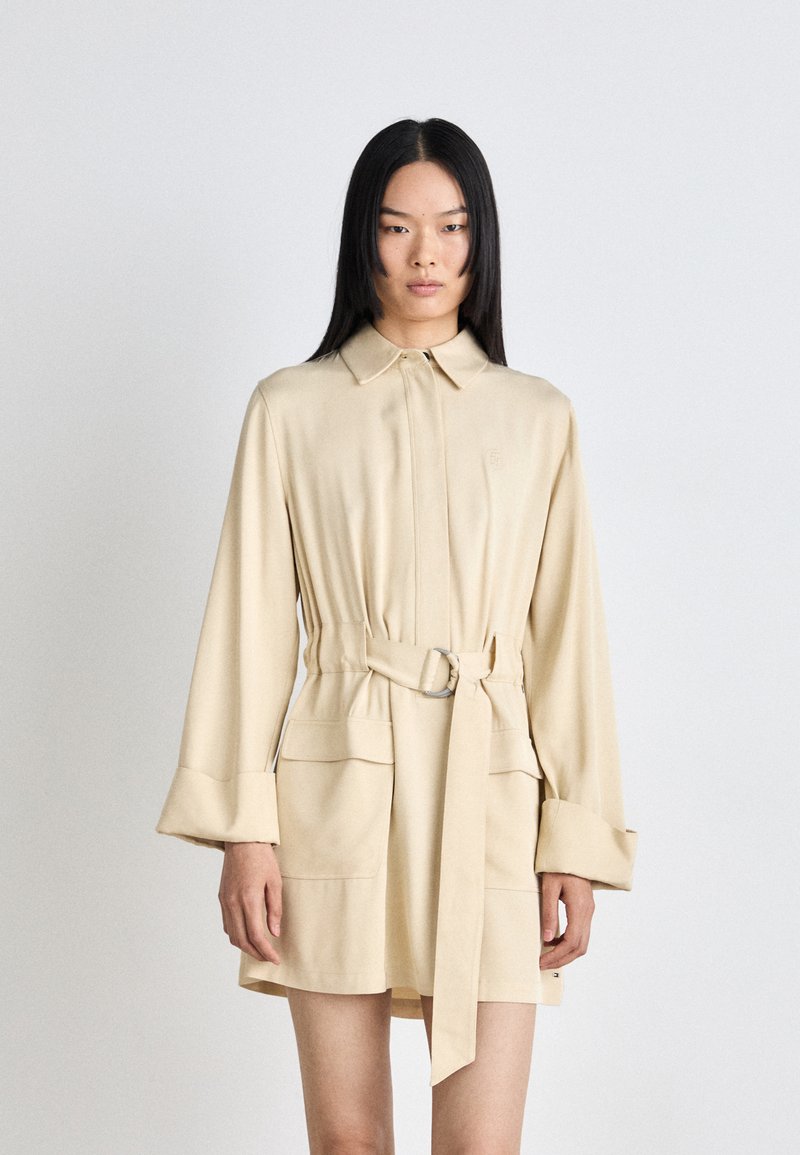 Béžové šaty s opaskom, golierom, dlhými rukávmi a prednými vreckami. Tkanina pôsobí hladko s miernym leskom. Jednoduchý dizajn a prispôsobený strih.
