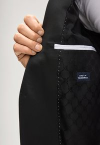 Interno di giacca nera con tessuto liscio e motivo geometrico. Include una piccola tasca bianca cucita e un'etichetta "STRETCH TECHNOWOOL".