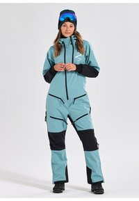 Een teal en zwarte skikostuum met een ritssluiting aan de voorkant, meerdere zakken en een comfortabele pasvorm. Voorzien van een hoge kraag en verstelbare manchetten.