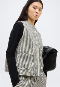 Νεαρή γυναίκα που φοράει μαύρη μπλούζα με μακρύ μανίκι, αμάνικο εντυπωσιακό γιλέκο με ριγέ ασπρόμαυρα σχέδια και ταιριαστό παντελόνι, κρατώντας μια μαύρη δερμάτινη τσάντα.