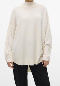 Personne portant un pull en tricot crème oversize à manches longues avec des poignets côtelés, associé à un pantalon noir, sur un fond blanc.