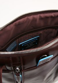 Borsa in pelle marrone con zip argentata, caratterizzata da una superficie liscia e tasche interne. Etichetta blu ben visibile con il marchio all'interno.