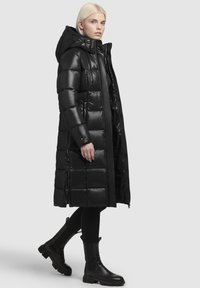 Black padded puffer coat with a hood, featuring a glossy finish, zippered side pockets, and a long silhouette, paired with black ankle boots.